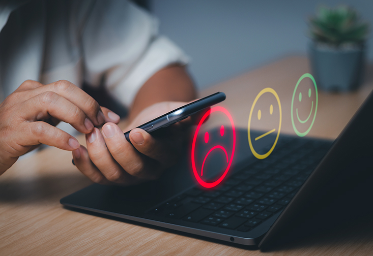 Close up on hands typing on a cell phone. Three emoticons showing sad, disappointed, and happy emotions.