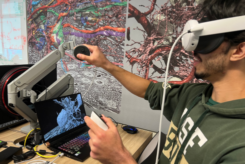 Student wearing VR goggles holds controllers in hands in front of computer with images of neurons