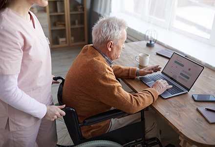 An older adult in a wheelchair is using a laptop at a wooden table, with a healthcare worker in pink scrubs standing nearby, offering assistance. The setting appears to be a home or care facility, with a warm and supportive atmosphere.